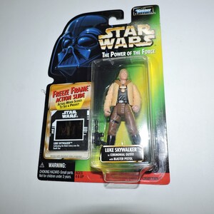 Op de afbeelding: Een Star Wars-actiefiguur van Luke Skywalker in een bruine ceremoniële outfit met een blasterpistool. De figuur is verpakt in een plastic blisterverpakking met een groene achtergrond en de tekst "Star Wars: The Power of the Force" op de voorkant.