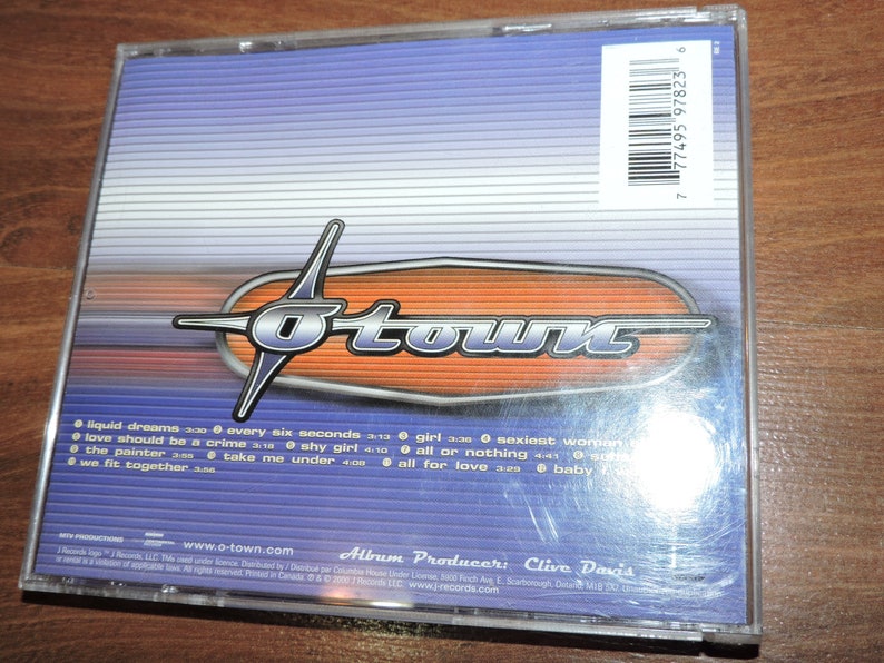 May include: A blue and orange CD case with the band name "O-Town" printed in white. The back of the case lists the song titles "Liquid Dreams", "Every Six Seconds", "Love Should Be a Crime", "The Painter", "Shy Girl", "Take Me Under", "Girl", "All or Nothing", "All for Love", and "Baby" with their corresponding lengths. The album producer is listed as "Clive Davis".