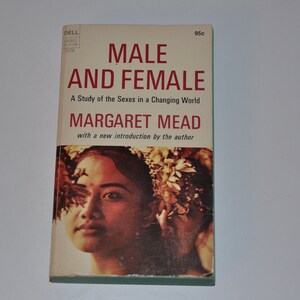 以下が含まれることがあります： 赤いタイトル「MALE AND FEMALE」と著者名「MARGARET MEAD」が書かれたビンテージのペーパーバックの表紙。表紙には、花を髪につけた女性の画像が掲載されています。