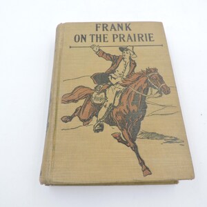 May include: A vintage hardcover book titled "FRANK ON THE PRAIRIE." The cover shows a brown and tan illustration of a man on horseback, with the title in black letters above. The book has a tan cover.