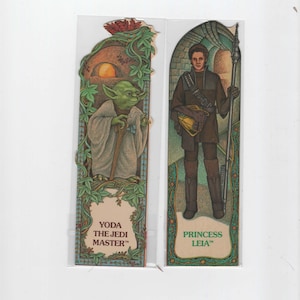 May include: Two Star Wars bookmark cards. The left card features Yoda, the Jedi Master, in a green robe with a brown walking stick. The right card features Princess Leia in a brown tunic and trousers, holding a staff. Both cards have a green, blue, and gold border.