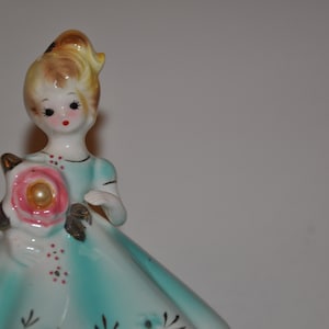 May include: A porcelain figurine of a girl in a blue dress holding a pink flower with a pearl centre. The girl has blonde hair and a painted face with red lips.