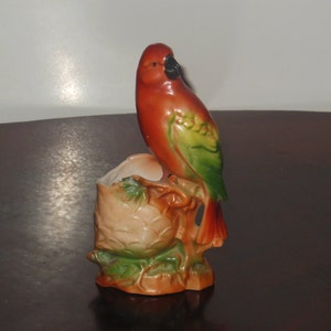 Könnte beinhalten: Eine Keramik-Vogelfigur mit einem roten, grünen und gelben Körper, die auf einem braunen und grünen Sockel sitzt. Der Vogel hat einen schwarzen Schnabel und schwarze Augen.