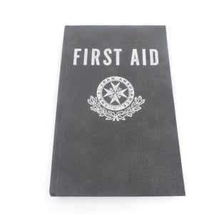 May include: A black first aid manual with the words "FIRST AID" in white block letters. The St. John Ambulance emblem is centered below the text. The book is rectangular and sits on a white surface.