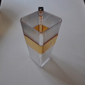 May include: A clear glass bottle with a gold band and a silver top. The bottle is empty and has a textured surface.