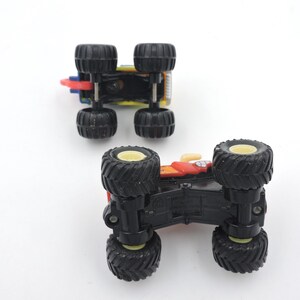 May include: Two toy monster trucks are shown from below, revealing their black tires and chassis. One truck is red, yellow, and blue, while the other is red and yellow. The tires have a black tread and yellow rims.