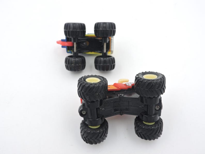 May include: Two toy monster trucks are shown from below, revealing their black tires and chassis. One truck is red, yellow, and blue, while the other is red and yellow. The tires have a black tread and yellow rims.