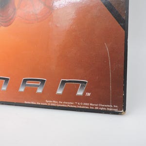 Op de afbeelding: Close-up van een doos met het woord "MAN" in zilveren blokletters. De achtergrond is een verloop van oranje en bruin. De tekst onderaan luidt "Spider-Man" en copyrightinformatie.