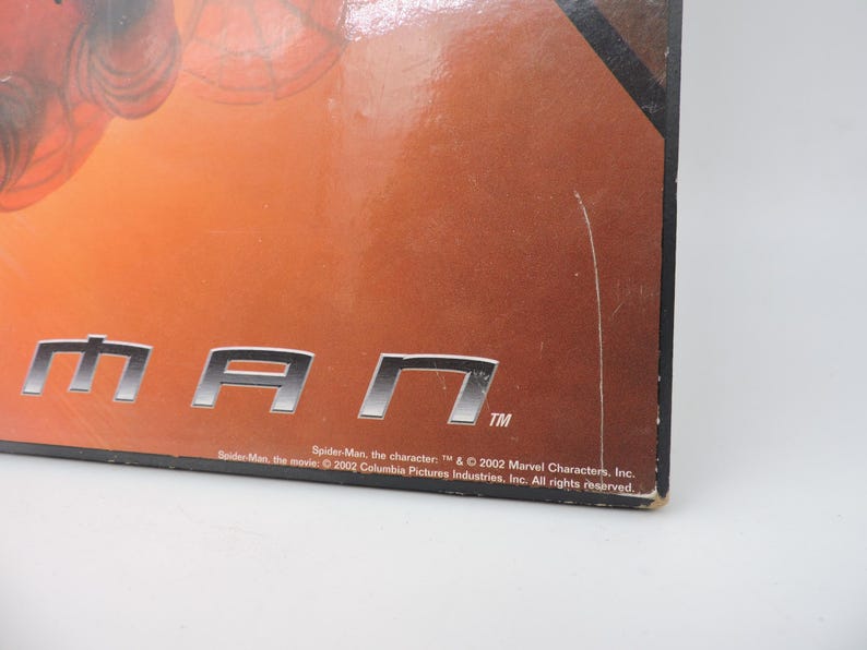 Op de afbeelding: Close-up van een doos met het woord "MAN" in zilveren blokletters. De achtergrond is een verloop van oranje en bruin. De tekst onderaan luidt "Spider-Man" en copyrightinformatie.