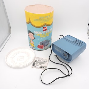 Könnte beinhalten: Ein blauer View-Master-Projektor mit einem schwarzen Kabel. Der Projektor steht auf einer weißen Oberfläche. Es gibt eine gelbe und rote Box mit einem Bild von Charlie Brown darauf. Die Box trägt die Aufschrift "View-Master Charlie Brown Theatre".