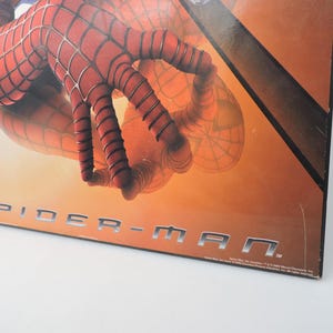 Op de afbeelding: Een close-up van een Spider-Man-afbeelding. De afbeelding toont de rood-zwarte gehandschoende hand van het personage met webdetails. De titel "SPIDER-MAN" wordt weergegeven in zilveren blokletters tegen een oranje achtergrond.
