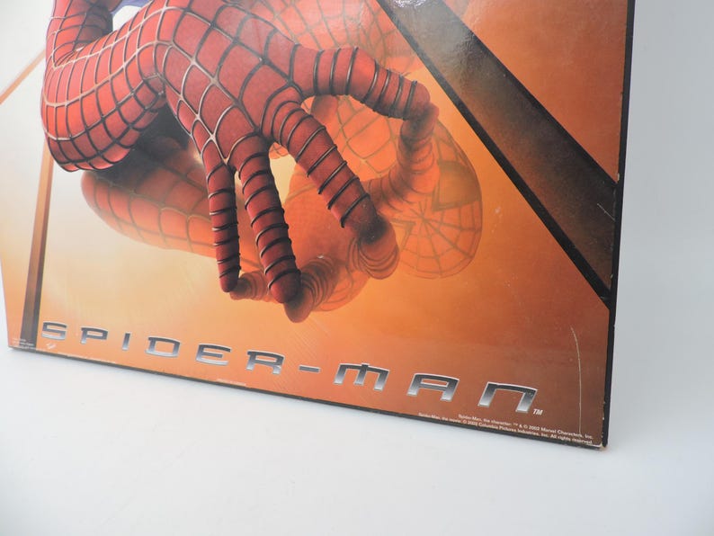 Op de afbeelding: Een close-up van een Spider-Man-afbeelding. De afbeelding toont de rood-zwarte gehandschoende hand van het personage met webdetails. De titel "SPIDER-MAN" wordt weergegeven in zilveren blokletters tegen een oranje achtergrond.