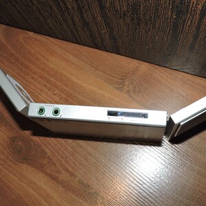 May include: A silver-colored portable audio device with a fold-out design. The device has two headphone jacks labeled "PHONES" A and B, and a small LCD screen.