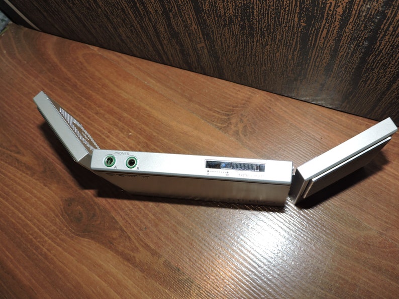 May include: A silver-colored portable audio device with a fold-out design. The device has two headphone jacks labeled "PHONES" A and B, and a small LCD screen.