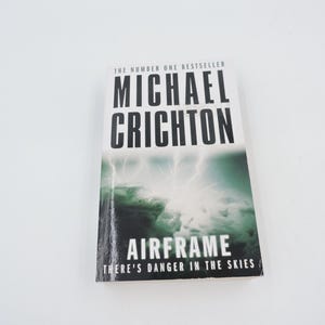 Può includere: Un libro tascabile intitolato "Airframe" di Michael Crichton. La copertina presenta un'immagine drammatica di fulmini su un paesaggio verde e grigio. È visibile anche il testo "THERE'S DANGER IN THE SKIES".