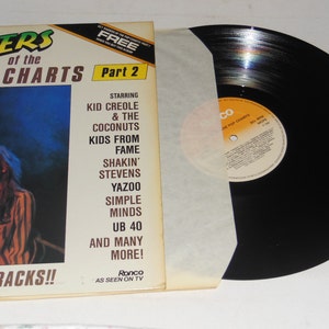 May include: A black vinyl record with a white paper inner sleeve. The record cover is yellow and orange and features a photo of two people. The text on the cover reads 'Riders of the Pop Charts Part 2', 'Starring Kid Creole & The Coconuts, Kids From Fame, Shakin' Stevens, Yazoo, Simple Minds, UB 40, And Many More!' and 'Hitting Tracks!!' The text on the record label reads 'Ronco, As Seen On TV, The Pop Charts, Hits & More'. 
