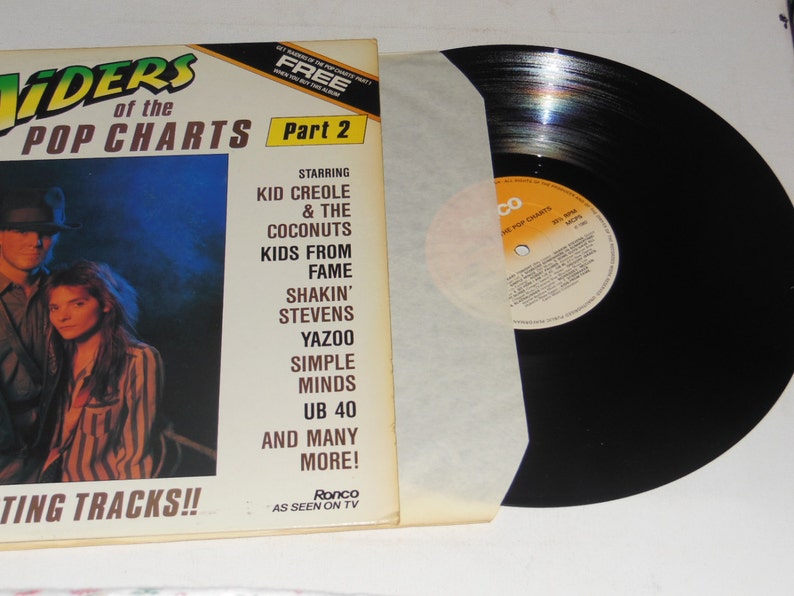 May include: A black vinyl record with a white paper inner sleeve. The record cover is yellow and orange and features a photo of two people. The text on the cover reads 'Riders of the Pop Charts Part 2', 'Starring Kid Creole & The Coconuts, Kids From Fame, Shakin' Stevens, Yazoo, Simple Minds, UB 40, And Many More!' and 'Hitting Tracks!!' The text on the record label reads 'Ronco, As Seen On TV, The Pop Charts, Hits & More'. 
