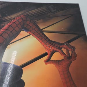 Op de afbeelding: Close-up van een Spider-Man-afbeelding. De afbeelding toont de rood-zwarte webben handen van het personage die een cirkelgebaar maken. De achtergrond toont een stadsgezicht en een warme, oranje getinte lucht.