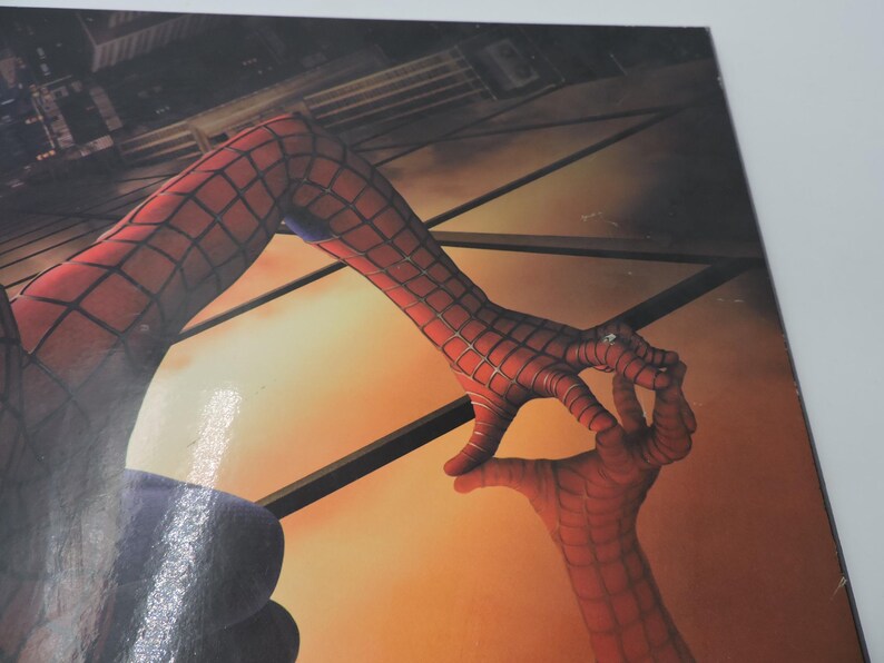 Op de afbeelding: Close-up van een Spider-Man-afbeelding. De afbeelding toont de rood-zwarte webben handen van het personage die een cirkelgebaar maken. De achtergrond toont een stadsgezicht en een warme, oranje getinte lucht.
