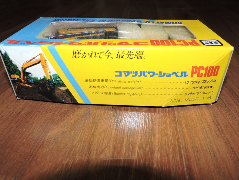 May include: A yellow toy Komatsu PC100 hydraulic excavator with a black cab and black tracks. The box is yellow and has Japanese text that reads "Komatsu Power Shovel PC100" and "Scale Model 1/48".
