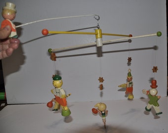Könnte beinhalten: Ein Holz-Babymobil mit fünf hängenden Figuren. Die Figuren sind in leuchtenden Farben bemalt, darunter Rot, Gelb, Grün, Blau und Weiß. Das Mobile verfügt über einen Stern, einen Clown, einen Tiger, ein Mädchen und einen Jungen. Das Mobile ist an einer weißen Holzstange aufgehängt.