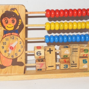 May include: Wooden toy abacus with a monkey face and clock on one side and colourful beads on the other. The abacus has wooden blocks with numbers and maths symbols. The numbers on the blocks are 5, 6, 4, 7, 9, 11, 12, 7, 3, 5, 4. The maths symbols are plus, minus, and equals.