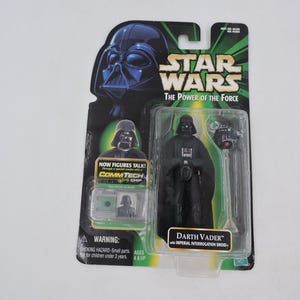 May include: A Star Wars Darth Vader action figure in its original packaging. The packaging features the Star Wars logo, the text "The Power of the Force", and a warning label. The figure is black and comes with an Imperial Interrogation Droid.
