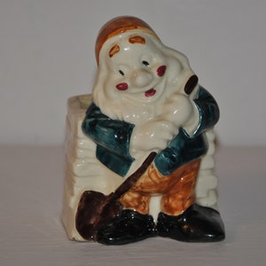 May include: A ceramic gnome figurine holding a shovel, wearing a blue jacket, brown trousers, and a brown hat. The gnome is sitting on a white planter.