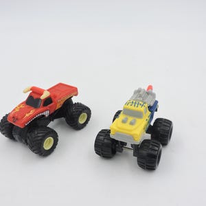 May include: Two toy monster trucks on a white background. One is red with a bull design, the other is yellow and blue. Both have large black tires and are designed for play.