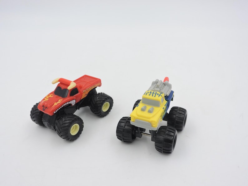 May include: Two toy monster trucks on a white background. One is red with a bull design, the other is yellow and blue. Both have large black tires and are designed for play.