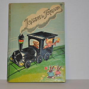 Peut inclure: Une couverture de livre pour enfants vintage avec un train jouet bleu et noir sur une voie verte. Le titre du livre est "Joanne in Toyland" avec un nuage blanc derrière le titre. Deux lapins sont au premier plan.