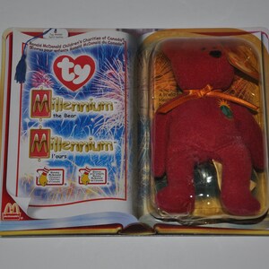 May include: A red Ty Beanie Baby bear called Millennium, with a gold ribbon and a green and gold tag, is packaged in a clear plastic case with a colourful cardboard backing. The backing features the Ty logo and the words "Millennium the Bear" and "Millennium l'ours".