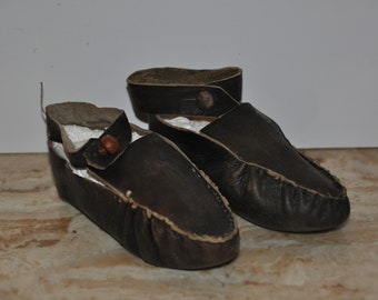 May include: A pair of brown leather moccasins with a strap and buckle. The moccasins have a worn look and are made of a soft leather.