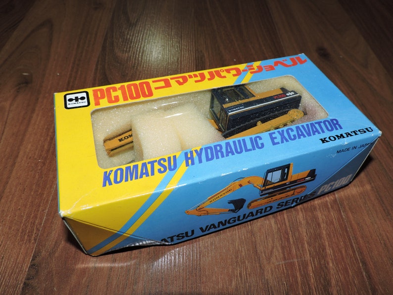 May include: A yellow and black toy Komatsu PC100 hydraulic excavator in a yellow and blue box with the text "PC100 コマツパワーショベル KOMATSU HYDRAULIC EXCAVATOR KOMATSU MADE IN JAPAN ATSU VANGUARD SERIES"
