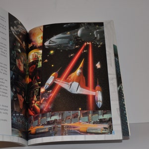 May include: A close-up of a book page with a picture of a spaceship in space. The spaceship is orange and white and has red laser beams coming from it. The text on the page reads "Anakin was streaking across the deck in his N-1 starfighter."
