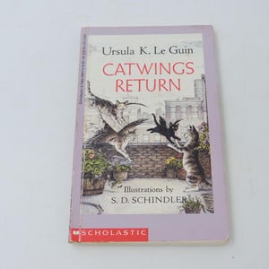 Könnte beinhalten: Ein Taschenbuch mit dem Titel "Catwings Return" von Ursula K. Le Guin. Das Cover zeigt Illustrationen von Katzen mit Flügeln, eine Stadtlandschaft und das Scholastic-Logo. Das Buch hat einen hellvioletten Rand.