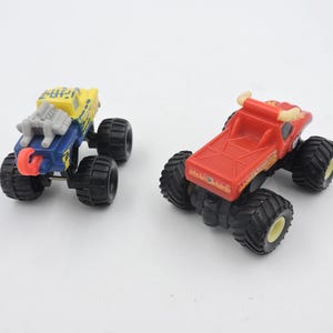 May include: Two toy monster trucks. One is blue and yellow with a silver engine and red accents. The other is red with yellow accents. Both have large black tires and are on a white background.