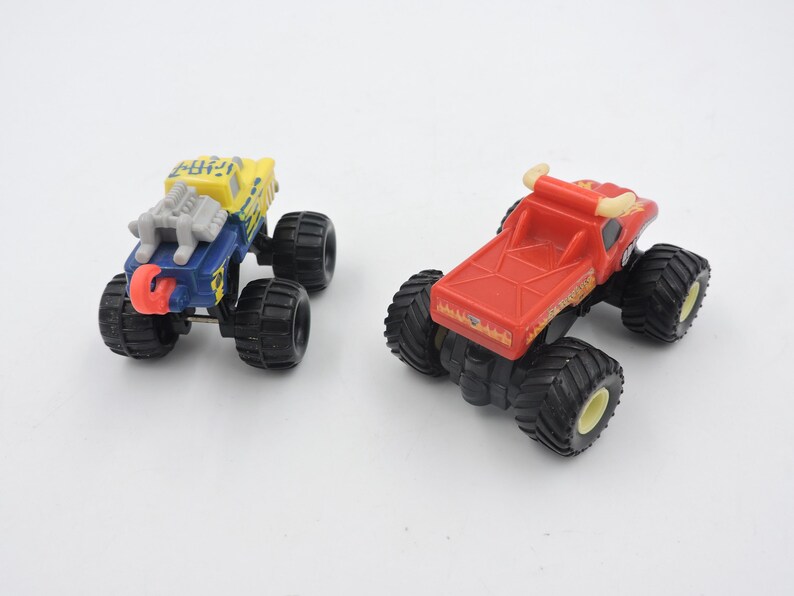 May include: Two toy monster trucks. One is blue and yellow with a silver engine and red accents. The other is red with yellow accents. Both have large black tires and are on a white background.