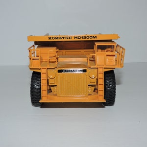May include: A yellow toy dump truck with the text "KOMATSU HD1200M" on the side. The truck has black tires and a black grill.