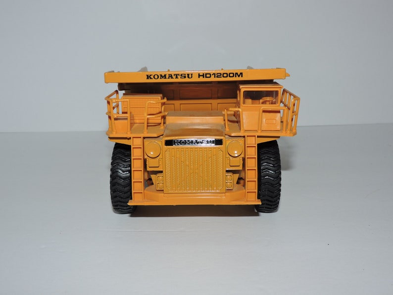 May include: A yellow toy dump truck with the text "KOMATSU HD1200M" on the side. The truck has black tires and a black grill.
