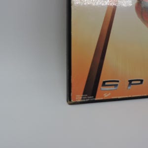 Op de afbeelding: Close-up van een doos met het woord "SPID" in zilveren letters. De achtergrond heeft een verloop van oranje en geel. De doos heeft een zwarte rand en staat op een wit oppervlak.