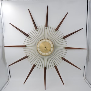 Puede incluir: Un reloj de pared moderno de mediados de siglo con un diseño de rayos de sol. El reloj presenta un centro dorado con una esfera crema y manecillas doradas. El reloj tiene largas púas de madera marrón que irradian hacia afuera, aproximadamente 76 cm de diámetro.