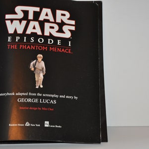 May include: A black book cover with the title "Star Wars Episode I The Phantom Menace." The cover features a young boy in a brown robe holding a bag. The book is by George Lucas.