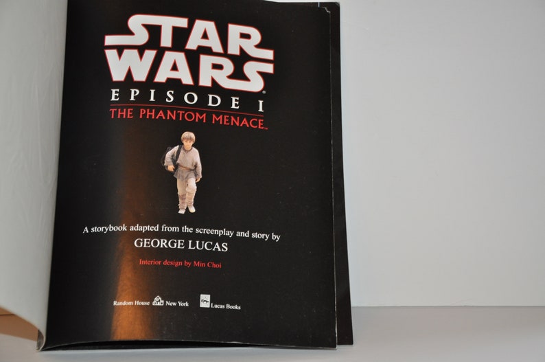 May include: A black book cover with the title "Star Wars Episode I The Phantom Menace." The cover features a young boy in a brown robe holding a bag. The book is by George Lucas.