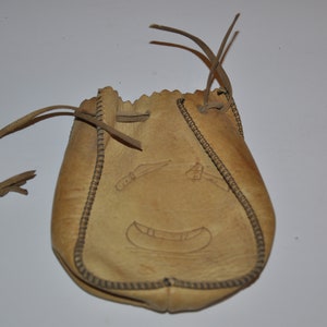 May include: A tan leather pouch with a drawstring closure and a decorative design of a canoe and two paddles. The pouch is stitched with a brown thread.