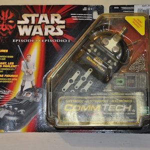 Puede incluir: Un juguete electrónico Star Wars Episodio I: Episodio I CommTech de color negro y dorado. El juguete presenta un pequeño dispositivo de mano negro con una antena plateada y un botón plateado. El empaque incluye una figura de un caballero Jedi con una espada láser.