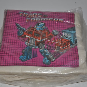 May include: A package of Transformers napkins with a pink background and a grid pattern. The image features Optimus Prime and Megatron in their robot forms. The text "TRANS FORMERS" is printed in white letters on the napkins.