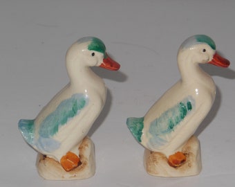 May include: A pair of ceramic duck salt and pepper shakers. The ducks are white with blue and green accents. They are standing on a base with a brown and orange design.