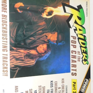 May include: A record album cover with a jungle scene. The album cover is titled 'Raiders of the Pop Charts Part 2.' The album cover features a man and a woman standing in front of a burning torch. The album cover lists the following artists: Kid Creole & The Coconuts, Kids From Fame, Shakin' Stevens, Yazoo, Simple Minds, UB 40, and many more. The album cover also includes the text '15 More Blockbusting Tracks!!' and 'Ronco Presents'. The album cover is in a black and white color scheme.