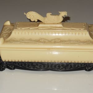 May include: A white ceramic trinket box with a black base. The lid features a raised design of a stylized bird with intricate details.